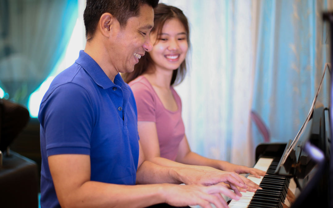 一名身穿藍色襯衫的男子在一名身穿粉紅色上衣的微笑的年輕女子旁邊彈鋼琴，兩人在藍色窗簾的房間裡享受音樂。