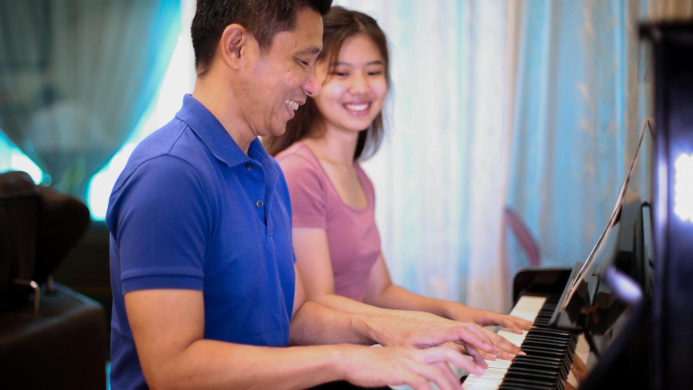 一名身穿藍色襯衫的男子在一名身穿粉紅色上衣的微笑的年輕女子旁邊彈鋼琴，兩人在藍色窗簾的房間裡享受音樂。
