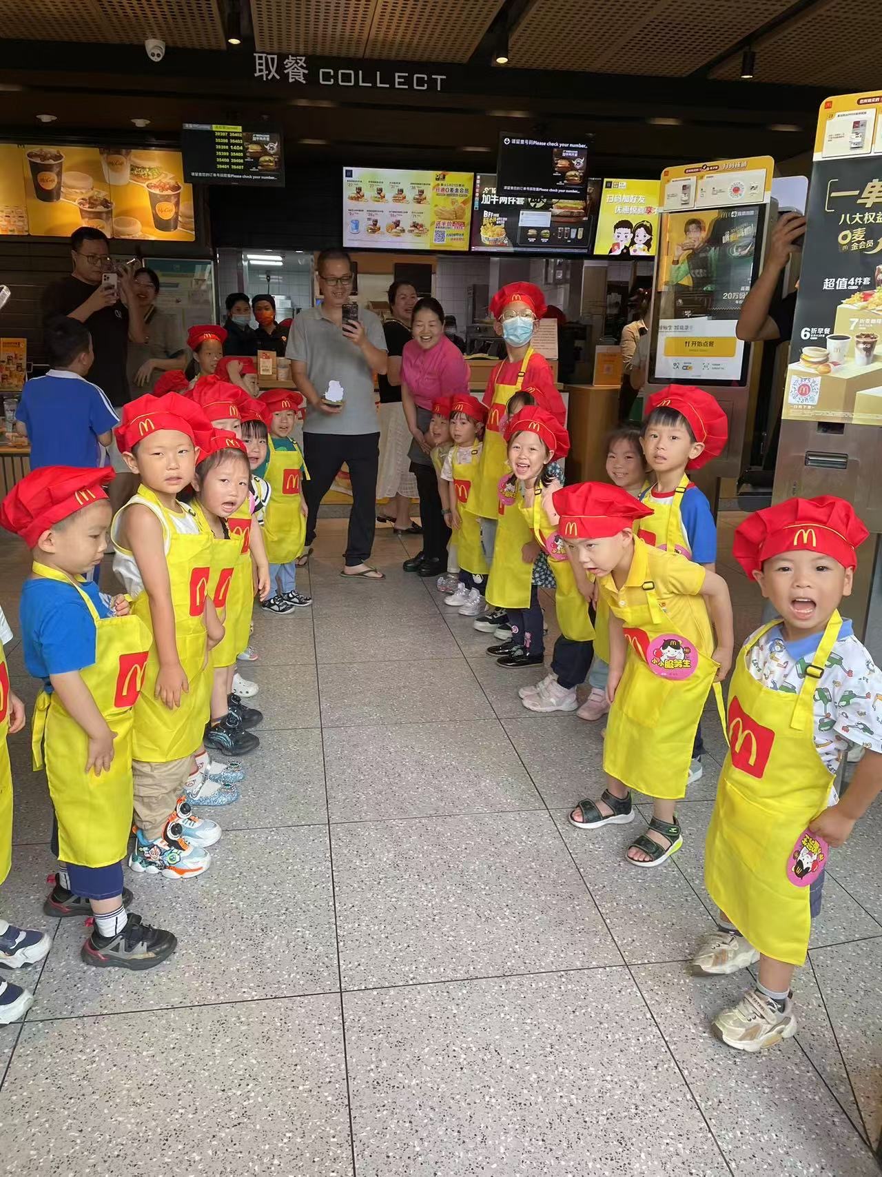 一群穿著黃色麥當勞圍裙、戴著紅色帽子的孩子在麥當勞餐廳內排成兩行，由成年人引導。