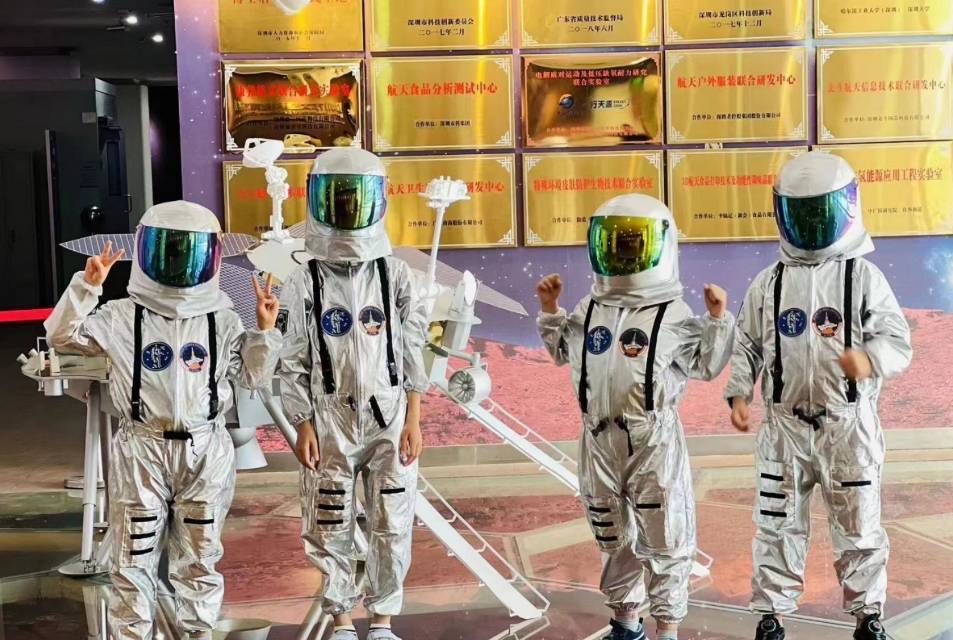 四名穿著反光太空人服裝的孩子站在室內，背景上以太空為主題，背景上展示著牌匾和太空船模型。