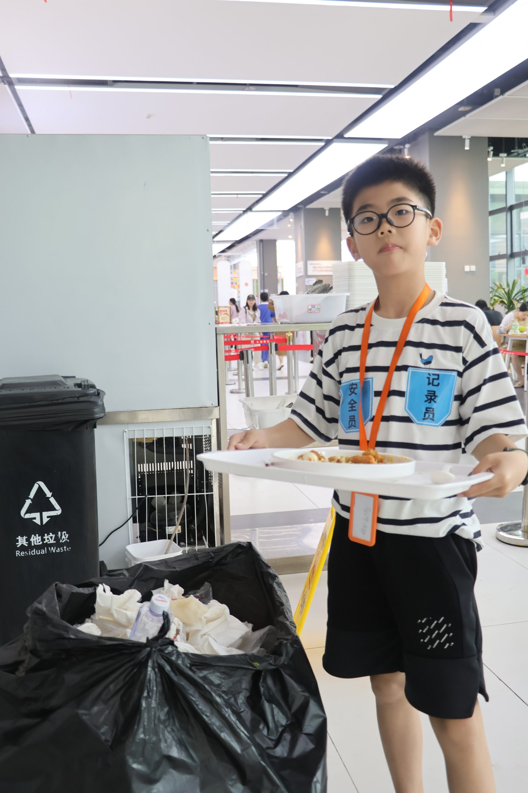 在一個類似自助餐廳的空間裡，一個戴著眼鏡、拿著托盤的男孩站在裝滿廢紙的垃圾桶旁。他穿著條紋襯衫、黑色短褲和橙色掛繩。