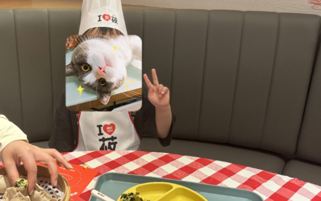 一個戴著貓面具、戴著寫著「我❤️貓」的廚師帽的孩子坐在一張桌子旁，桌子上放著一盤分開的食物和一個竹蒸籠。孩子用一隻手做出和平手勢。