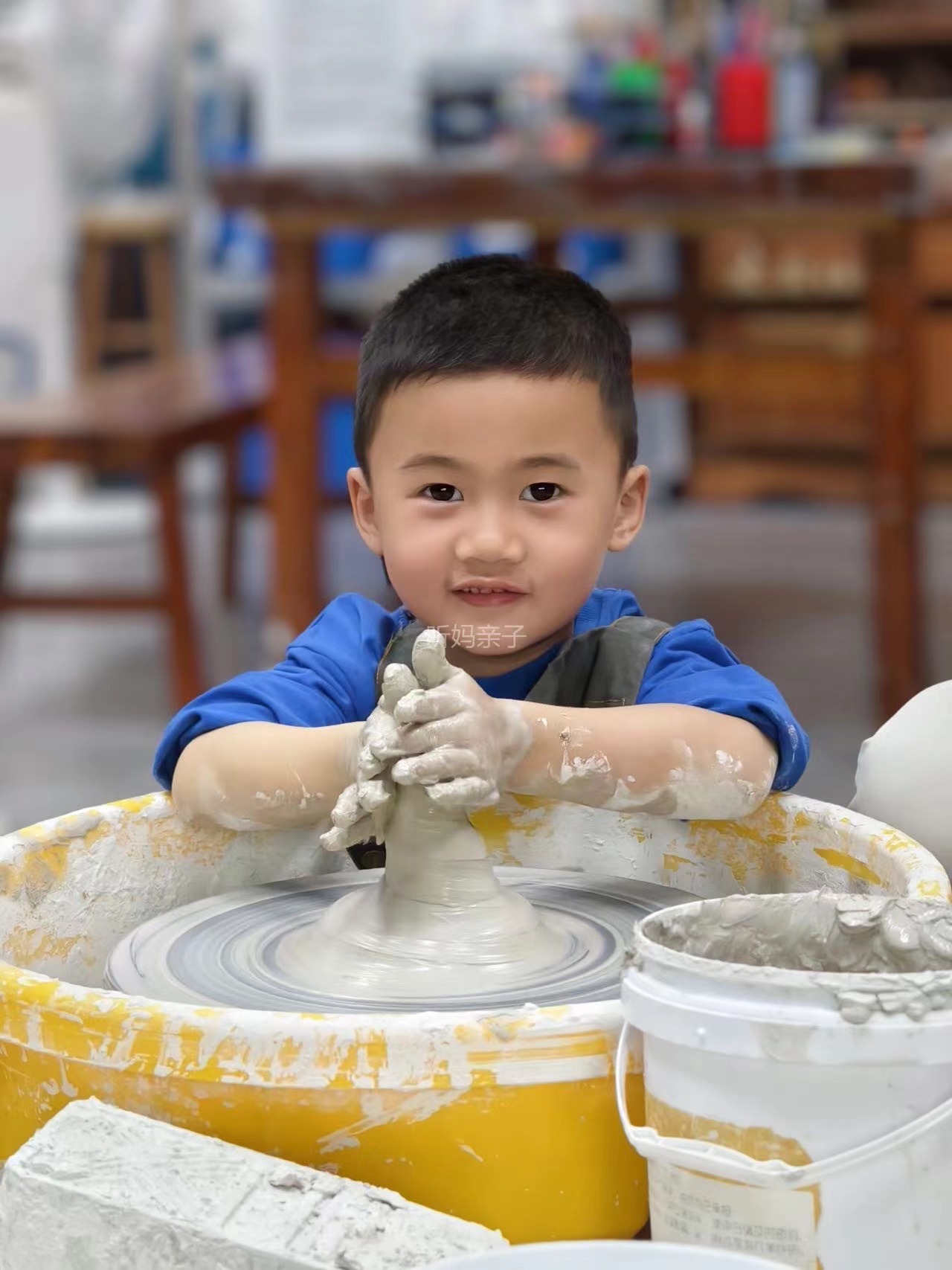 一個小孩正在陶輪上專心地塑造黏土。他的手上沾滿了濕黏土，背景中可以看到各種陶器和材料。