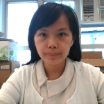 一位黑色直髮的女士坐在室內的辦公桌前,望著鏡頭。背景可見紙箱和冷氣機。.