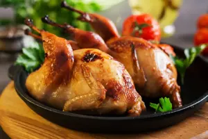 在黑色平底鍋中盛上兩隻烤鹌鹑,配上新鮮歐芹,背景是番茄和油菜。.