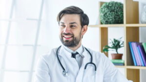 一位穿著白大褂、戴著聽診器的男醫生坐在明亮的辦公室裡微笑,背景是架子和植物。.