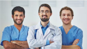 三位身穿藍色手術服和白大褂的男性醫療專業人員站在一起,在臨床環境中對著鏡頭微笑。.