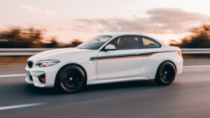 日落時分,一輛帶有賽車條紋的白色 BMW 轎跑車在高速公路上行駛,背景為動態模糊影像。.