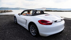 一個人坐在停在水體附近的白色敞篷跑車裡,背景是群山和多雲的天空。.