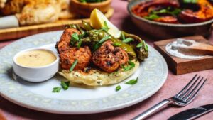 一盤烤肉串放在扁麵包上，配上烤青椒、檸檬片、小碗中的醬汁，以及盤子旁的刀叉。.