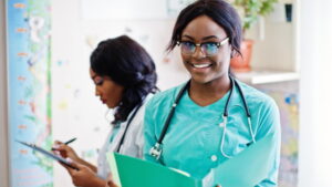 兩位穿著手術服的專業醫護人員站在醫療環境中,其中一位拿著文件夾對著鏡頭微笑,另一位則在背景的剪貼板上寫字。.