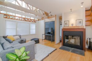 現代化客廳,天花板橫樑外露、灰色組合沙發、壁爐、電視、木地板、地毯,角落還有一盆大植物。.