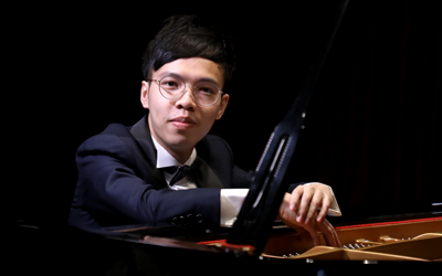 一位穿著西裝、戴著眼鏡的男子坐在三角鋼琴前，一隻手放在琴鍵上望著鏡頭。.