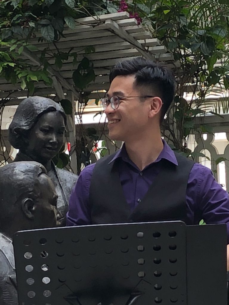 一位身穿紫色襯衫和黑色背心的男子站在音樂架前微笑，背景是兩座青銅雕像和綠色植物。.