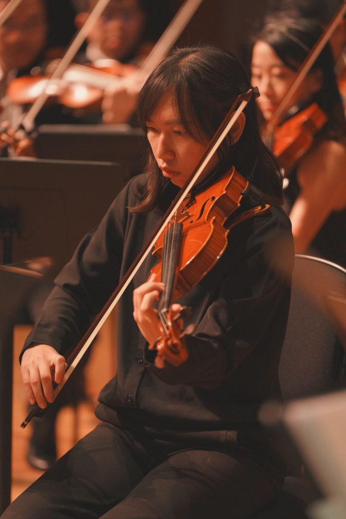 一位身穿黑色衣服的女士在管弦樂隊中拉小提琴，背景中可見其他小提琴手。.