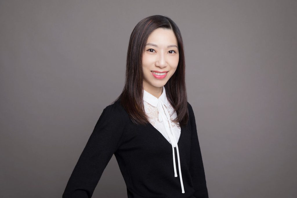 一位深色直髮、身穿黑色毛衣外罩白色上衣的女性，站在素淨的灰色背景中，對著鏡頭微笑。.